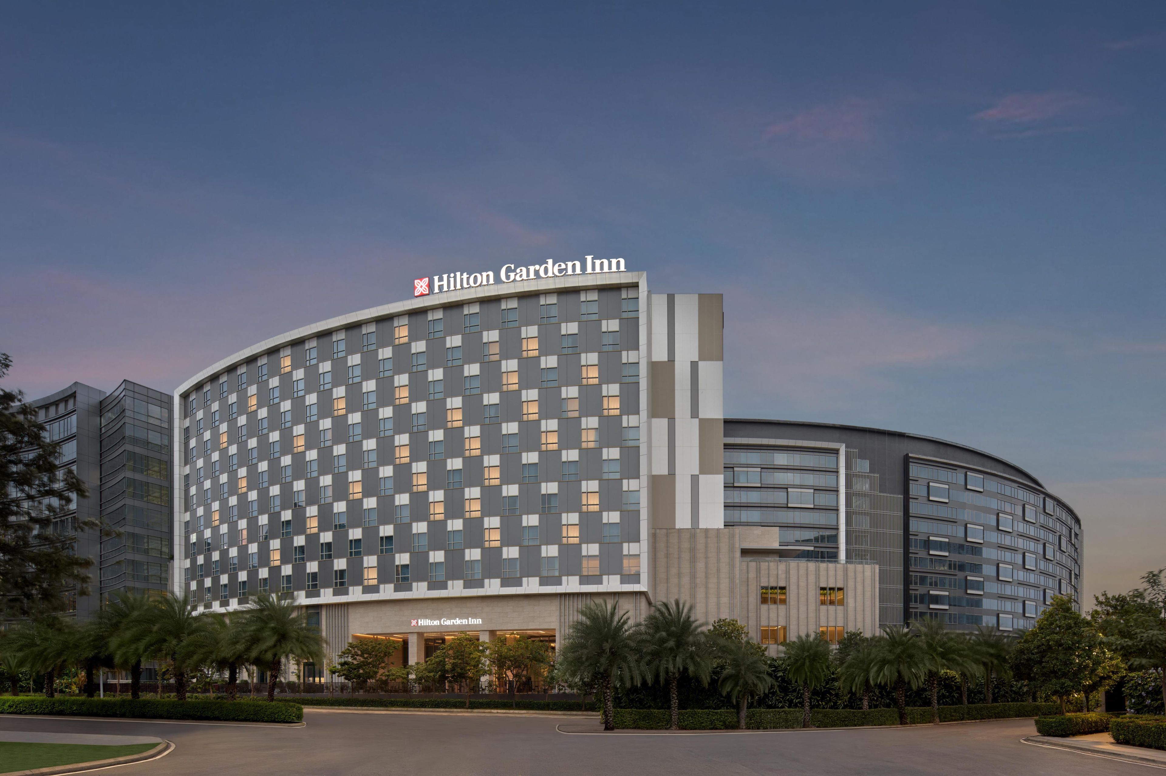 A modern Hilton Garden Inn hotel with a curved facade, large windows, and a well-maintained landscaped area with palm trees.