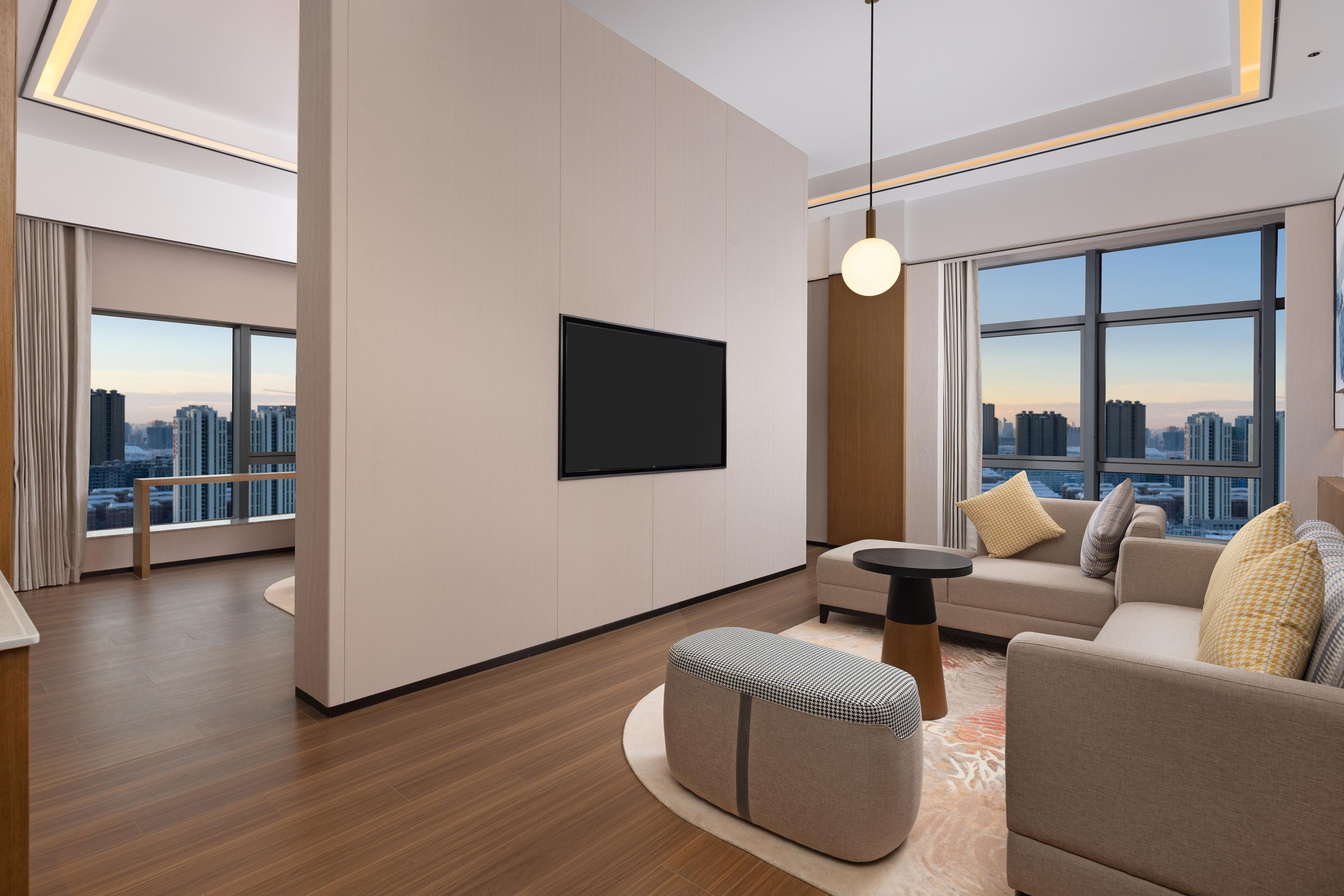 A modern living room with a flat-screen TV mounted on the wall, a sofa set, a round coffee table, and a large window offering a city view.