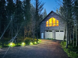 Eine Hütte mit einem großen Fenster, einer Garage und einer Auffahrt mit Schotter, umgeben von Bäumen.