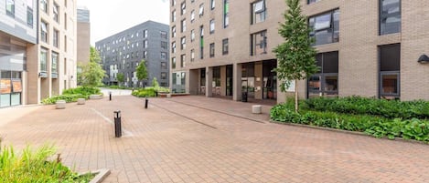 A modern building with multiple floors, large windows, and a paved courtyard with greenery.