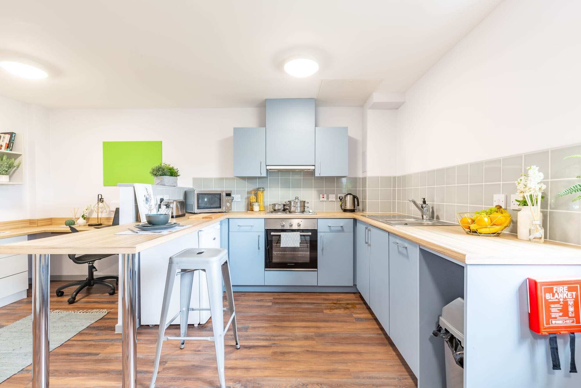 Eine moderne Küche mit hellblauen Schränken, einer Holz Arbeitsplatte, einem eingebauten Backofen und einer Mikrowelle. Ein Hocker und ein kleiner Tisch mit einer Schüssel und einer Pflanze sind ebenfalls vorhanden.