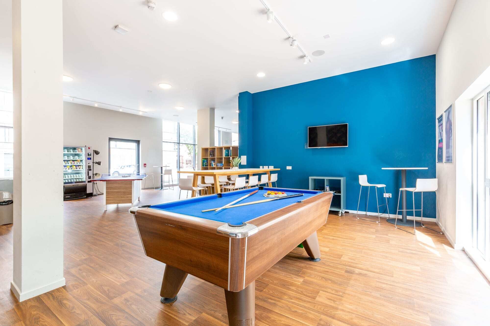 Una mesa de billar en una habitación bien iluminada con una pared azul de acento, una máquina expendedora y un área de descanso con mesas y sillas.