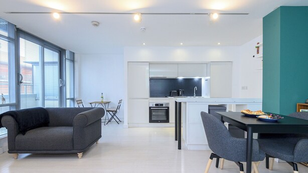 Una cocina moderna con una isla central, armarios blancos y un horno empotrado. Hay un comedor con mesa y sillas, y un sofá gris.