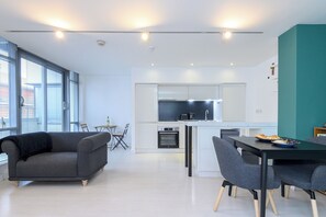 A modern kitchen with a central island, white cabinetry, and a built-in oven. There is a dining area with a table and chairs, and a gray sofa. - Stylish Central Retreat in the Heart of Leeds - Pass the Keys (Leeds)