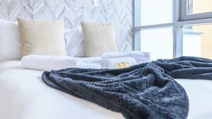 Una cama bien tendida con sábanas blancas y una manta gris oscuro, una ventana con vistas, y un papel tapiz con patrón de espiga.