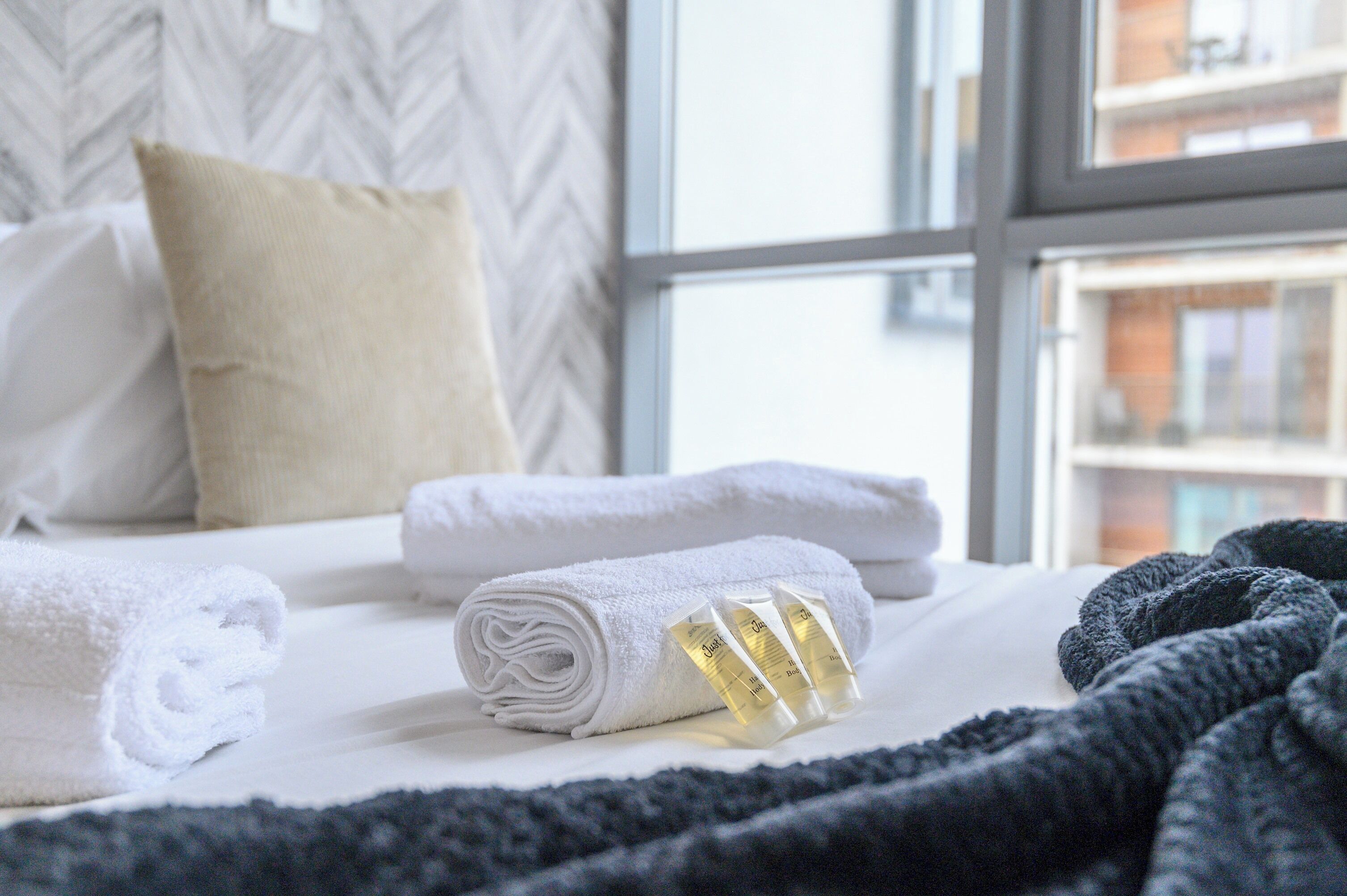A neatly made bed with rolled white towels and a black blanket, a beige pillow, and a window with a view of buildings.