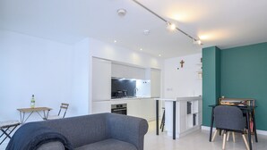 Una sala de estar moderna con un sofá gris, una pequeña mesa de comedor y una cocina con armarios blancos y encimera negra.