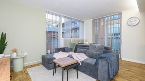 Ein modernes Wohnzimmer mit einer grauen Eckcouch, einem kleinen Holztisch und einem weißen Teppich. Links befindet sich eine Zimmerpflanze und ein Beistelltisch mit Dekorationsobjekten.