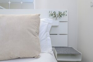 Un lit avec un oreiller beige et un oreiller blanc, une table de chevet blanche avec une plante, et une boîte de rangement grise.