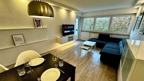 Een moderne woonkamer met een eetgedeelte, een zwarte bank, een televisie en grote ramen.