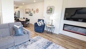Ein Wohnzimmer mit grauem Sofa, blauem Sessel, Kamin, Fernseher und Essbereich im Hintergrund.