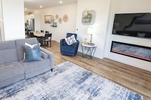 Een woonkamer met een grijze bank, een blauwe fauteuil, een open haard, een televisie en een eethoek op de achtergrond.