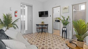 Un salon moderne avec un canapé, une table à manger et des chaises. Un téléviseur est fixé au mur, ainsi qu’un extincteur et un panneau indiquant la mention « REGARDEZ ».