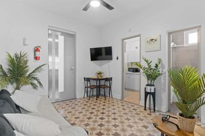 A modern living room with a sofa, a dining table, and chairs. There is a TV mounted on the wall, a fire extinguisher, and a sign that says "LOOK".