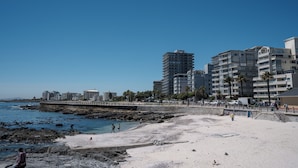 Een strand met een rotsachtige kustlijn, een steiger en moderne gebouwen op de achtergrond.