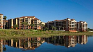 A multi-story hotel with a reflection in the water, surrounded by greenery and palm trees.