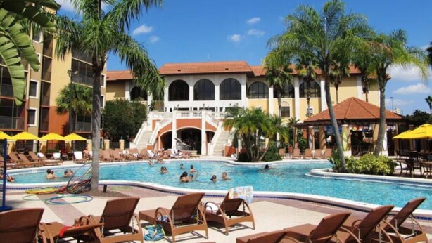 A pool area with lounge chairs, palm trees, and a building with arched windows.