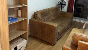 Ein Wohnzimmer mit einem braunen Ledersofa, einem Holzregal und einem Standventilator.