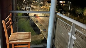 Ein Holzstuhl auf einem Balkon mit Metallgeländer und Blick auf eine Straße mit Autos und Gebäuden.