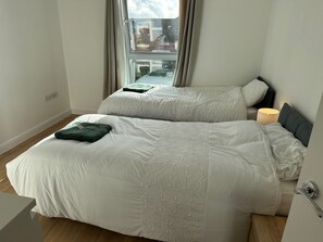 A room with two beds, a window with curtains, a bedside lamp, and a visible electrical outlet. - Staying With Ganmos Place (Bracknell)