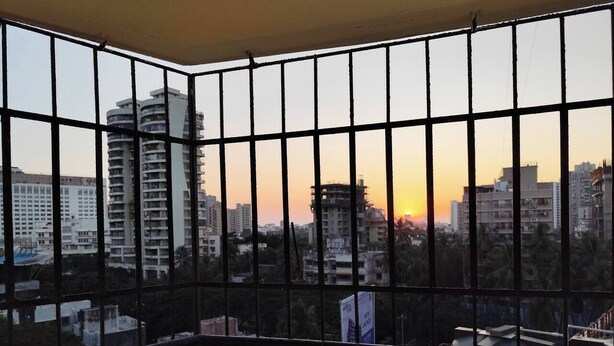 A view through a barred window with a sunset in the background, featuring multiple high-rise buildings and some construction sites. - Stylish 2BHK near Lokhandwala, Kokilaben & NESCO (Mumbai)
