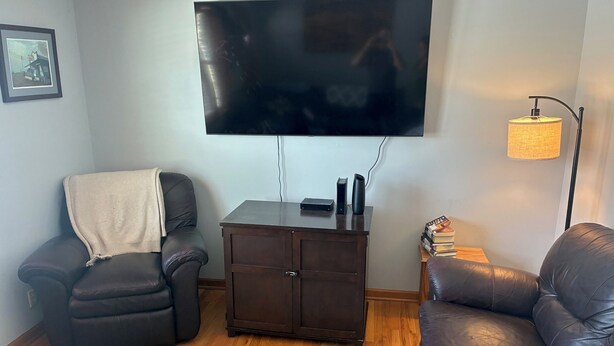 A living room with a leather sofa, a dark wooden cabinet, a TV mounted on the wall, and a lamp on a side table. - Kansas City World Cup Rental - PRICED RIGHT (Platte City)