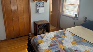 A bedroom with a wooden wardrobe, a small wooden table with books, a bed with a colorful quilt, a calendar on the wall, and a window with blinds. - Kansas City World Cup Rental - PRICED RIGHT (Platte City)