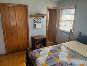 Ein Schlafzimmer mit einem Holzschrank, einem kleinen Holztisch mit Büchern, einem Bett mit farbenfroher Steppdecke, einem Kalender an der Wand und einem Fenster mit Jalousien.