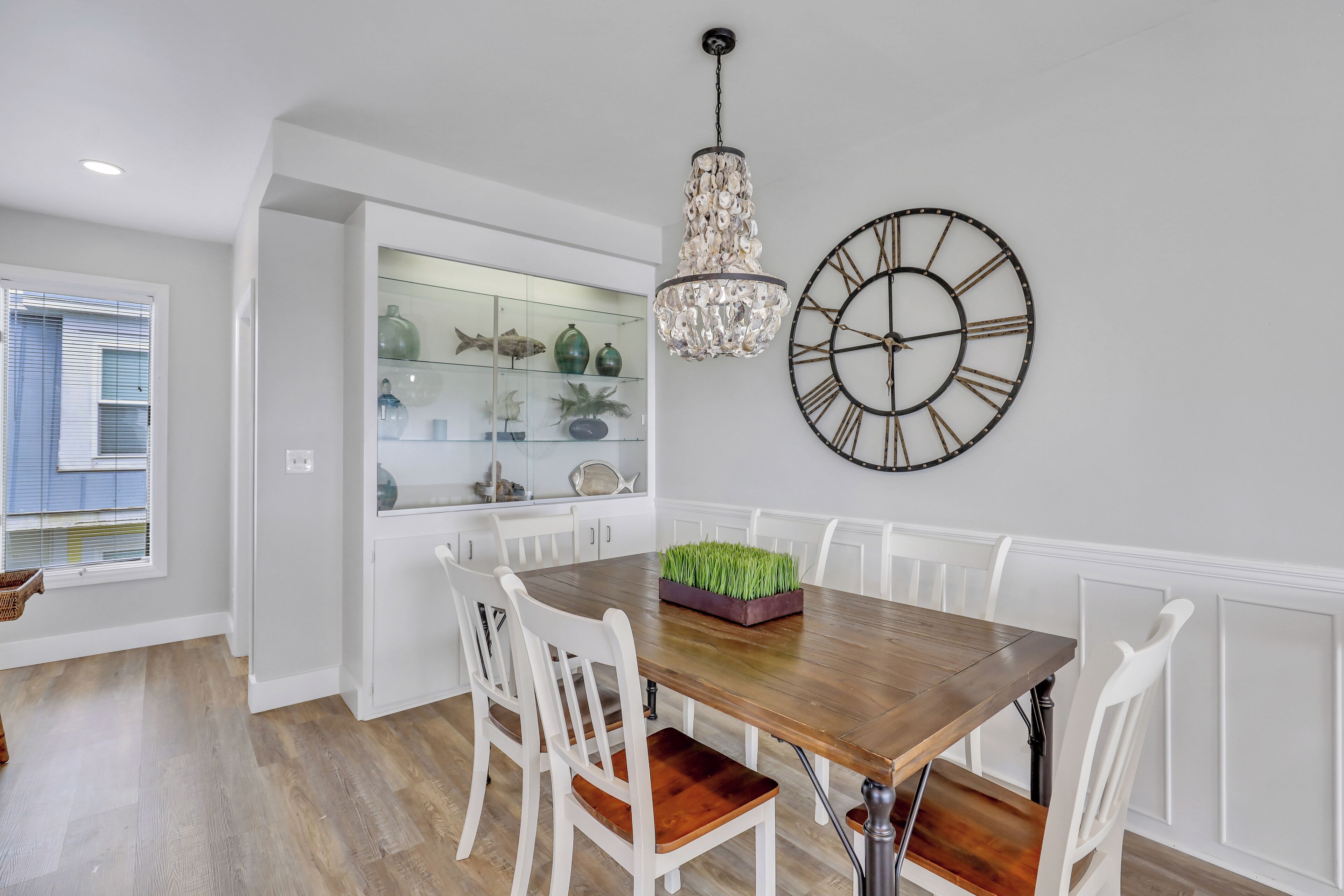 A dining area with a wooden table, white chairs, a chandelier, and a clock on the wall.