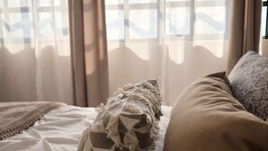 Ein Bett mit einer gemusterten Decke und einem braunen Kopfkissen, ein Fenster mit Bergblick und durchsichtigen Vorhängen.