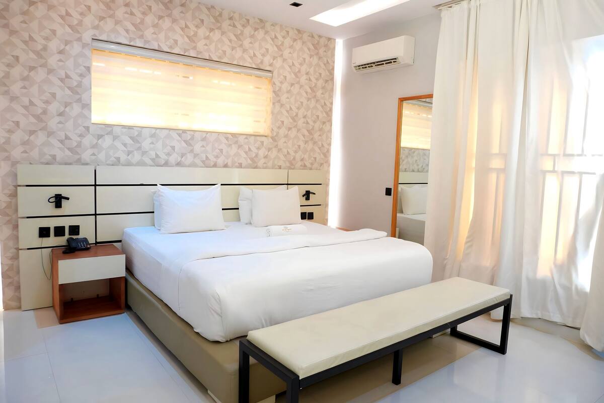 Une chambre à coucher bien aménagée, avec un lit, des tables de chevet, un banc, une fenêtre avec des stores et un appareil de climatisation.