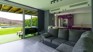 Ein modernes Wohnzimmer mit einem großen grauen Ecksofa, einem kleinen Tisch und einer Glastür, die auf eine Terrasse mit Pool und Grünflächen führt.