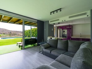 A modern living room with a large grey sectional sofa, a small table, and a glass door leading to a patio with a pool and greenery. - Salobre Golf Villas Dragos I (Maspalomas)