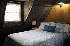 Un dormitorio con techo inclinado, cama con ropa de cama estampada, lámpara de noche y ventana con persianas.