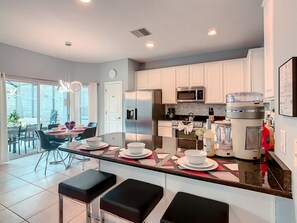 A modern kitchen with white cabinets, a stainless steel refrigerator, a microwave, and a coffee maker. There are two black bar stools and a dining table with chairs. - EPIC! Themed-Arcade-Theatre-Pool Table-Pool&Spa-3mi>Disney (Kissimmee)