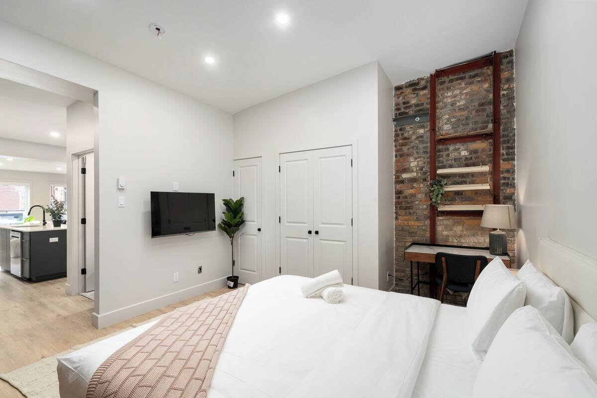A modern hotel room with a large bed, a desk, a TV, and a brick wall.