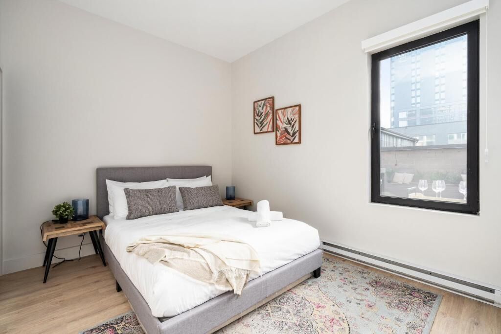 A modern bedroom with a large bed, a nightstand, a small table with a plant, and a window with a view of buildings.