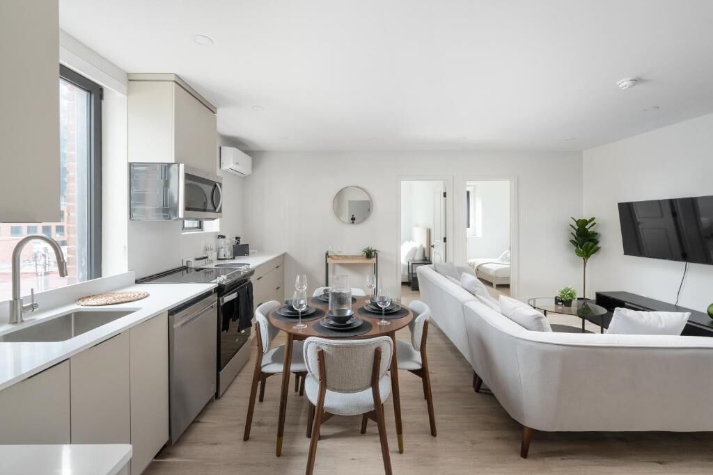 A modern kitchen with a dining area, a living room with a sofa, and a TV mounted on the wall.