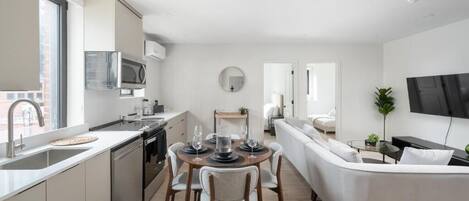 A modern kitchen with a dining area, a living room with a sofa, and a TV mounted on the wall.