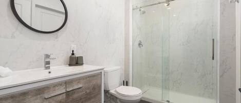 A modern bathroom with a marble wall, a wooden vanity, a white toilet, and a glass-enclosed shower.