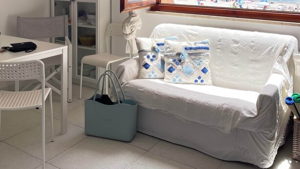 A room with a view of the beach, a white sofa, a table with chairs, and a cabinet. - THE WINDOW TO THE SEA (Alghero)