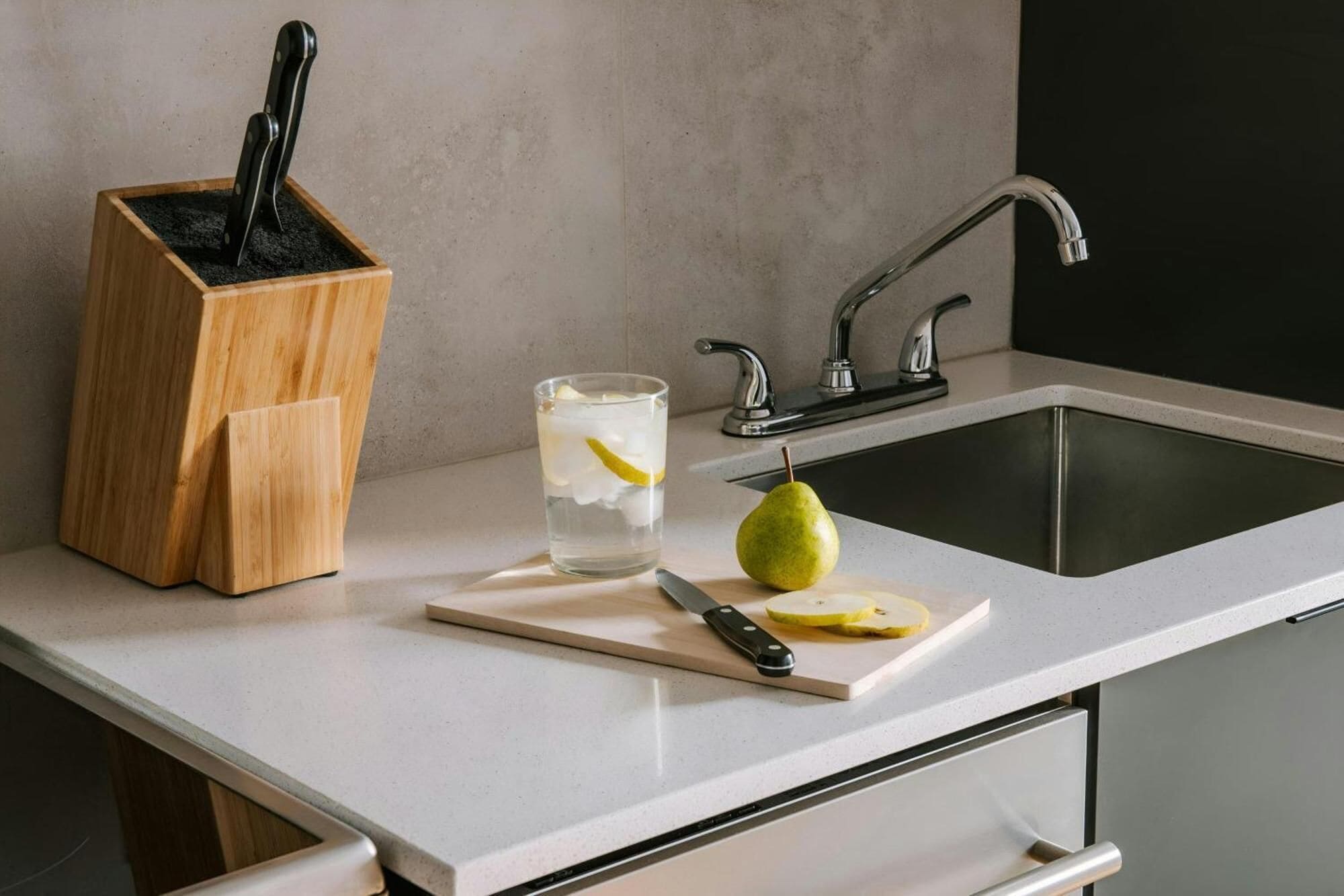 A kitchen counter with a knife block, a glass of water with a lemon slice, and a pear on a cutting board.