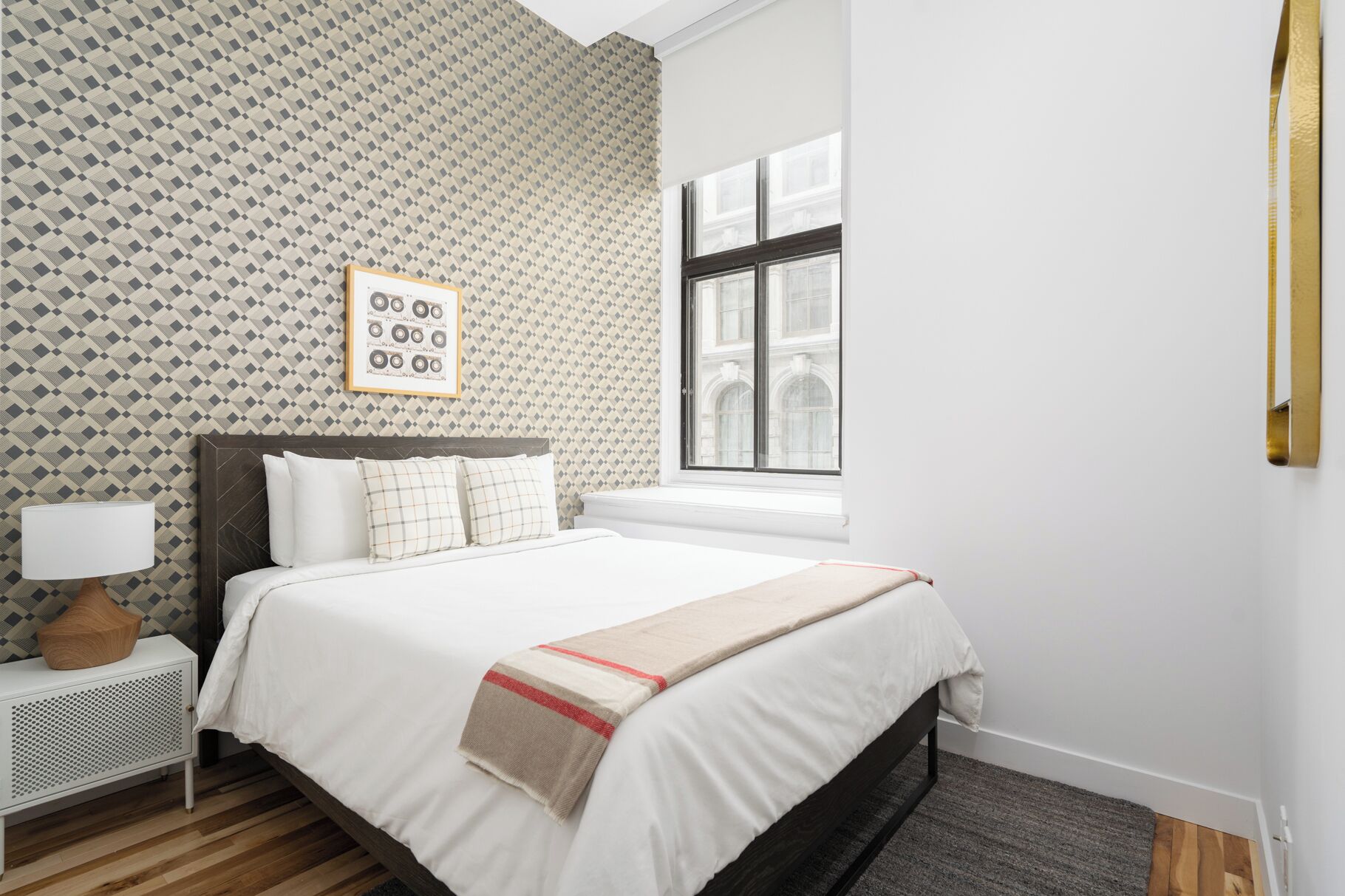 A bedroom with a patterned wallpaper, a bed with white bedding, a nightstand with a lamp, and a window with a view of buildings.