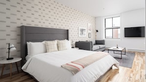A modern bedroom with a large bed, a grey sofa, a small table, and a TV mounted on the wall.
