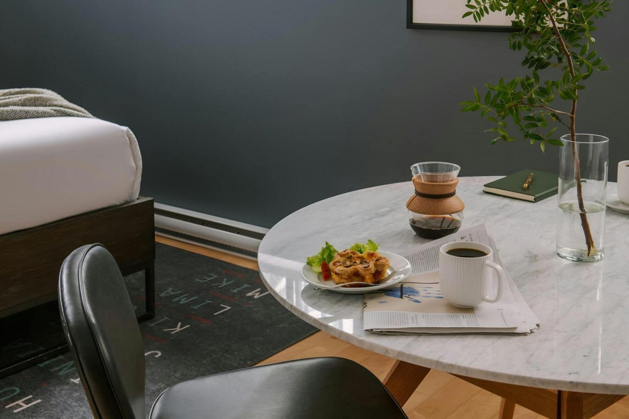 Una habitación moderna con una cama, una mesa redonda con un plato de comida, una taza de café, un vaso de agua, un libro y una planta.