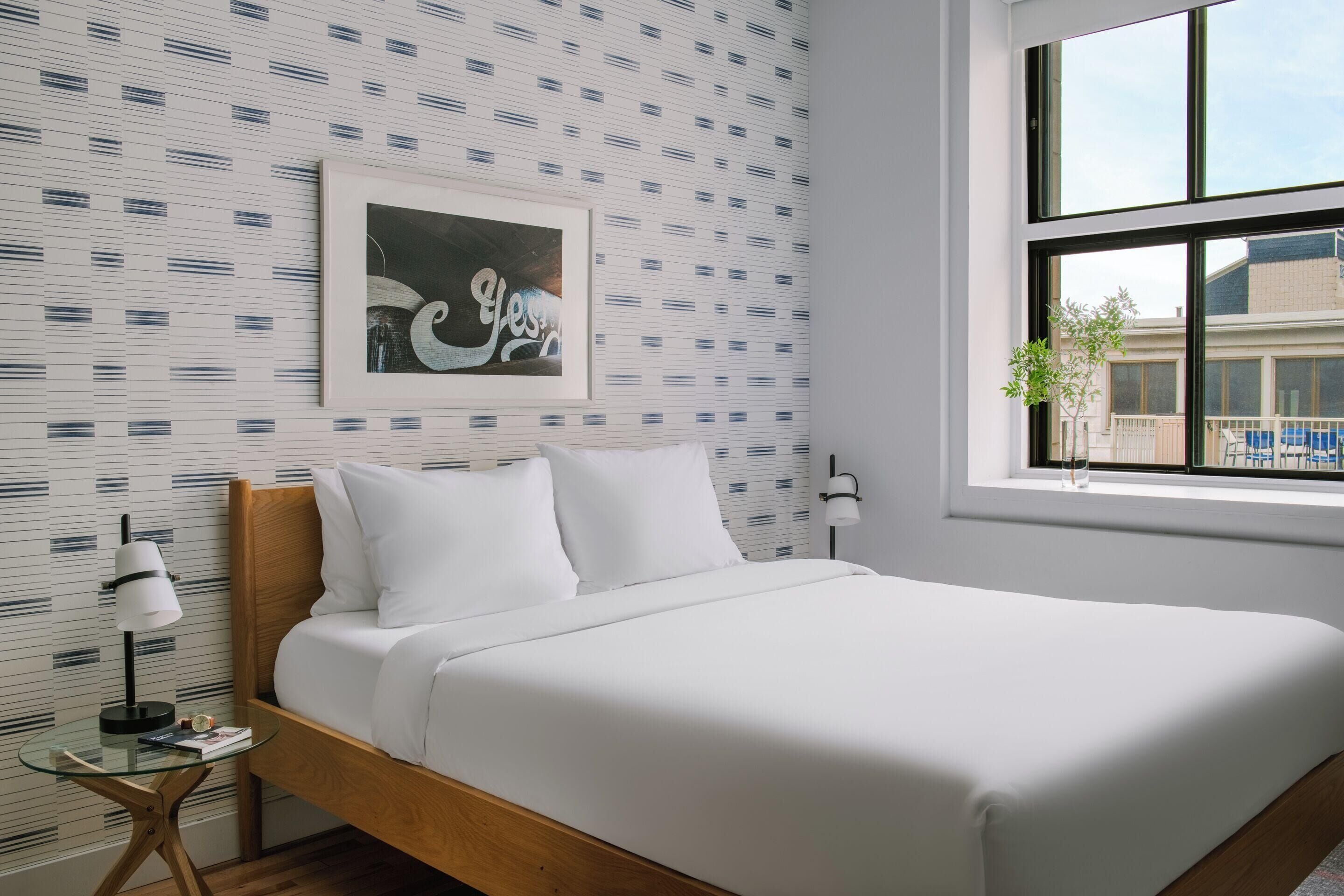 Un lit bien fait, avec du linge de lit blanc, un tableau encadré au mur et une fenêtre donnant sur des bâtiments à l’extérieur.