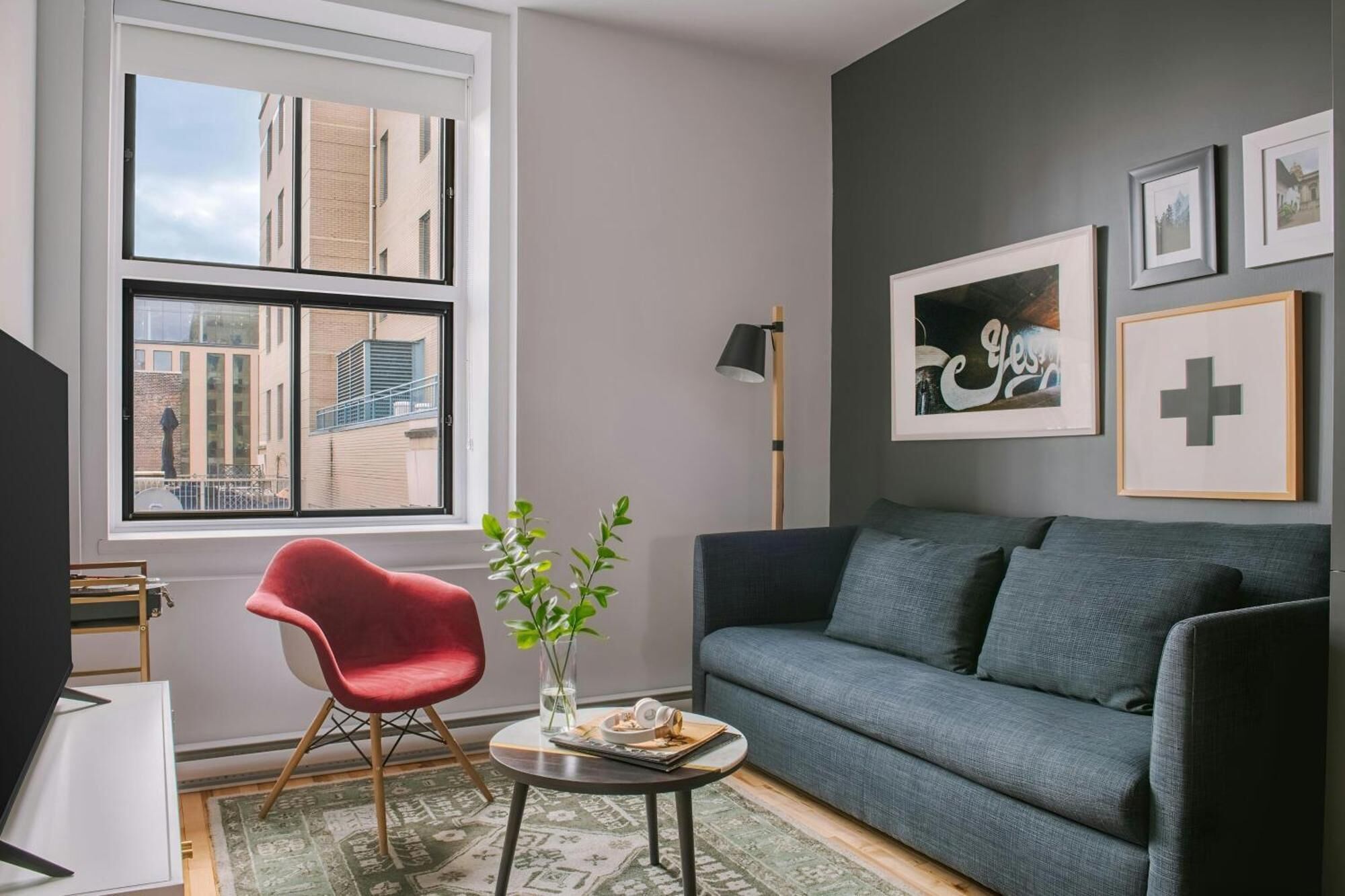 A modern living room with a grey sofa, a red chair, a small table with a plant, and a window with a city view.