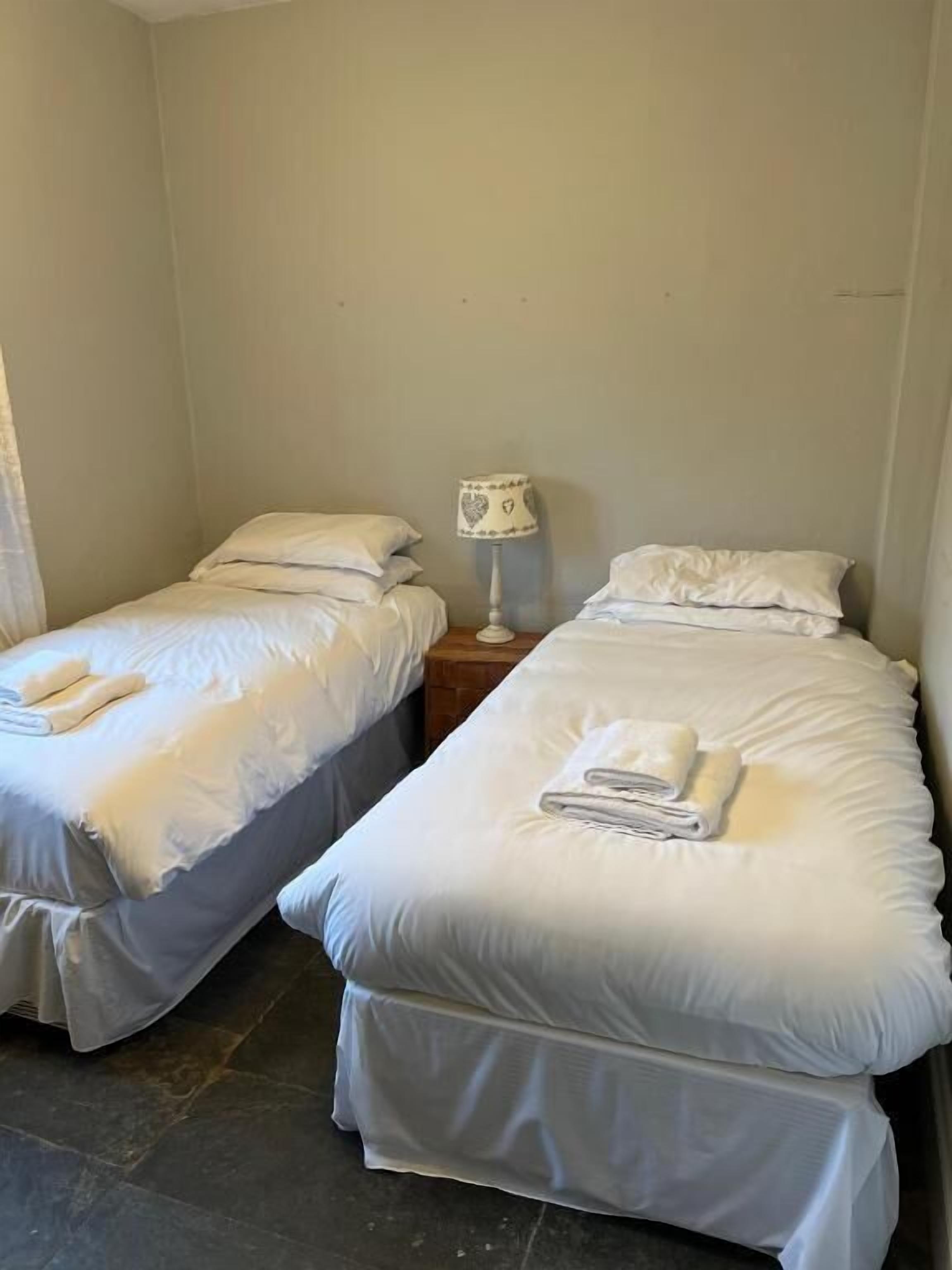 A small, clean room with two single beds, each with a folded towel and a bedside table with a lamp.