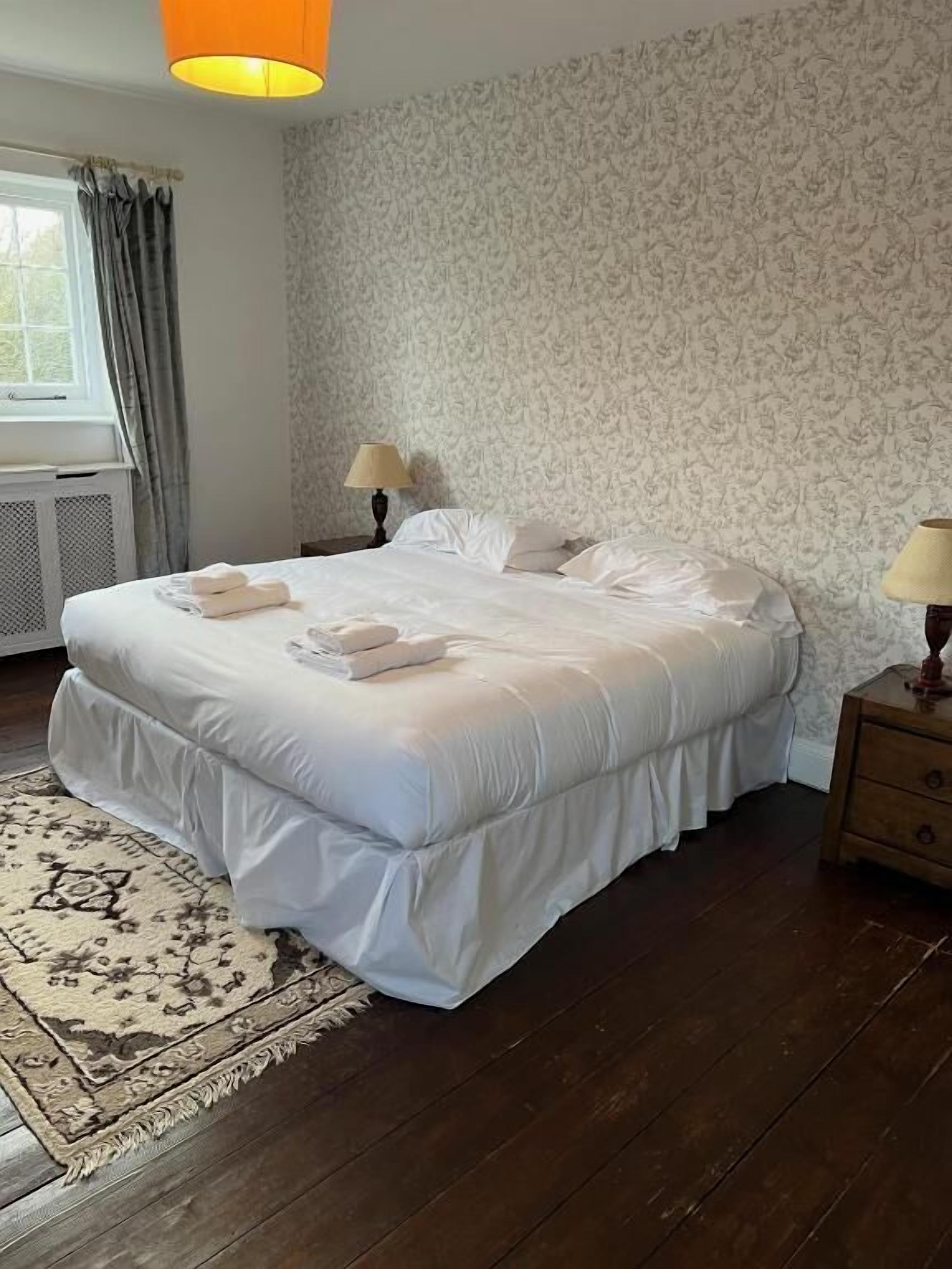 A neatly made bed with white linens and towels, a patterned rug, a wooden floor, a window with curtains, and a wall with a floral wallpaper pattern.