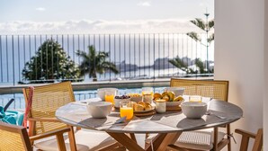 A round table with breakfast items, including bowls, glasses, and fruits, on a balcony overlooking a pool and the ocean. - Atlantic Bloom by Homie (Funchal)